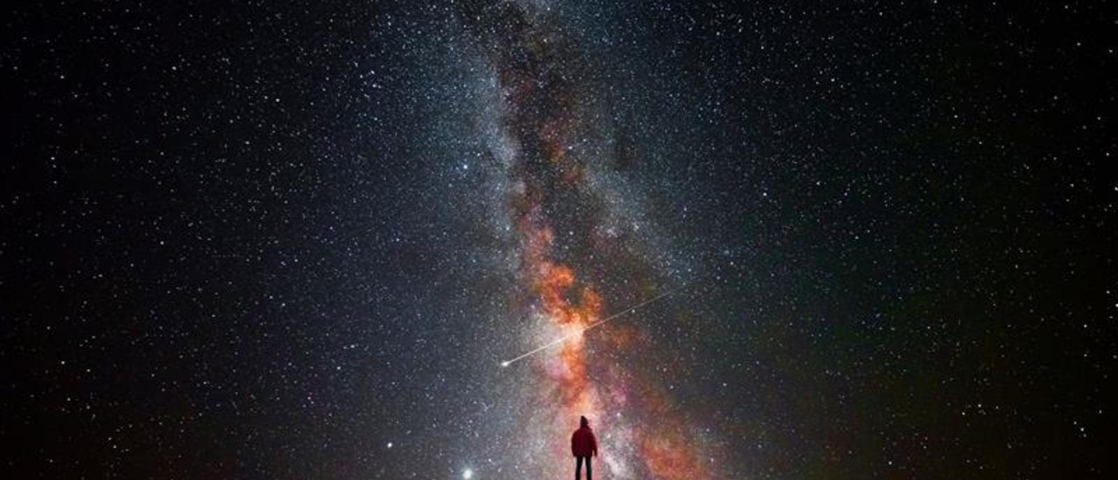 Céu estrelado em uma noite muito escura com iluminação média ao centro e uma pessoa em pé olhando pra cima 